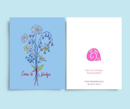 Front and back of greeting card. Right: Colour pencil drawing of bouquet of wild flowers tied with red bow. Daisy, buttercups, forget me nots, cowslips, barebells, red campion, bluebells, Handwritten text Caru ti blodyn. Left back @tytwtdesigns