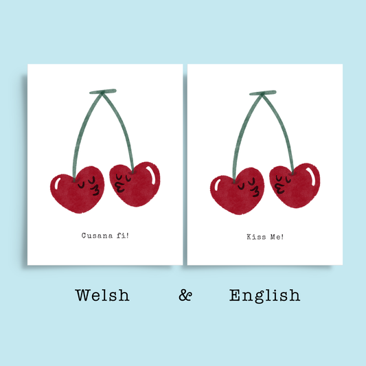 Romantic greeting card in Welsh and English. Hand drawn print of two cherries with faces kissing. Typewriter text below reads Cusana fi, Welsh, and Kiss me, English. 